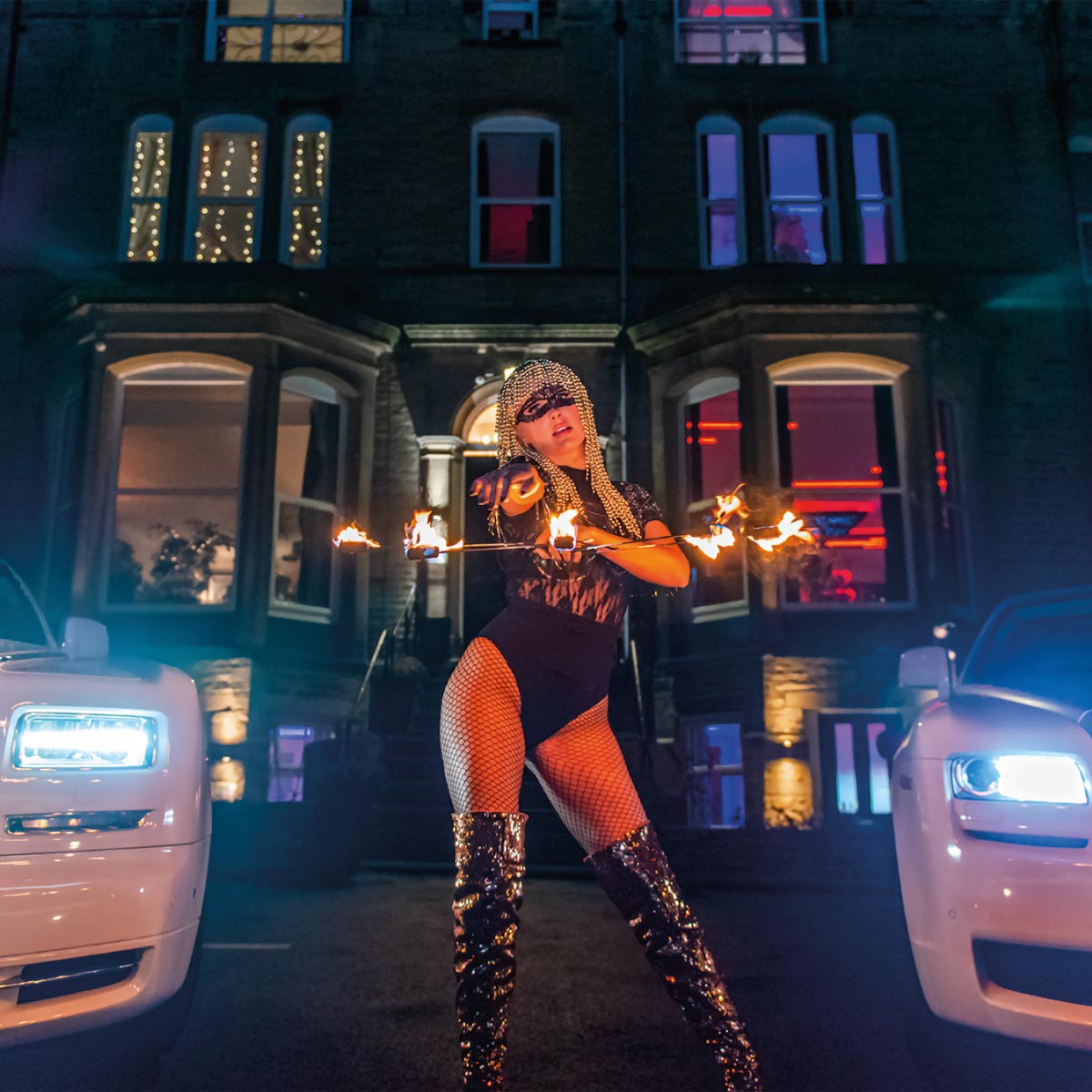 A female performer holding fire sticks next to a pair of Rolls Royce's.